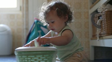 Kıvırcık saçlı çocuk mutfakta salata fırıldağıyla dikkatle oynuyor. Erken gelişim aşamasında yerde otururken sapını çevirmeye odaklanmış.