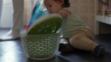 Kıvırcık saçlı meraklı çocuk, mutfak zemininde salata fırıldağıyla oynuyor, erken çocukluk gelişimi sırasında oturmuş ve araştırma yapıyor.