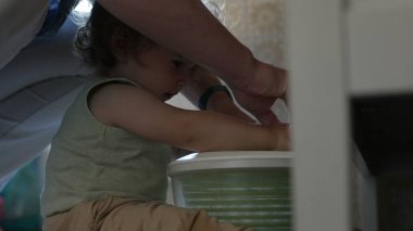 Kıvırcık saçlı çocuk, yetişkin elinin yardımıyla salata fırıldağıyla oynarken, oyun zamanındaki ilk keşifler sırasında etkileşim ve destek anı