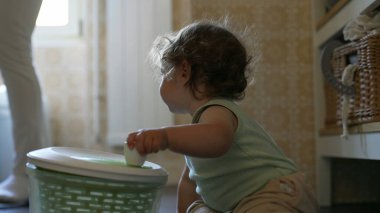 Kıvırcık saçlı çocuk mutfakta salata fırıldağıyla dikkatle oynuyor. Erken gelişim aşamasında yerde otururken sapını çevirmeye odaklanmış.
