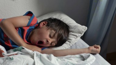 Yatakta uzandıktan sonra kollarını açarak uzanan genç çocuk, uykulu bir ifadeyle ve yumuşak yatak odası ışığında kıvırcık saçlarıyla yavaşça uyanıyor.
