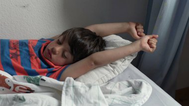 Yatakta uzandıktan sonra kollarını açarak uzanan genç çocuk, uykulu bir ifadeyle ve yumuşak yatak odası ışığında kıvırcık saçlarıyla yavaşça uyanıyor.