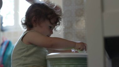 Kıvırcık saçlı çocuk, yetişkin elinin yardımıyla salata fırıldağıyla oynarken, oyun zamanındaki ilk keşifler sırasında etkileşim ve destek anı