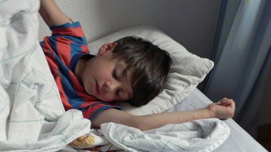 Yatakta uzandıktan sonra kollarını açarak uzanan genç çocuk, uykulu bir ifadeyle ve yumuşak yatak odası ışığında kıvırcık saçlarıyla yavaşça uyanıyor.
