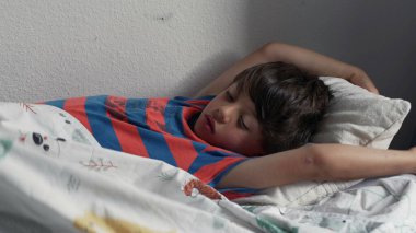 Yatakta uzandıktan sonra kollarını açarak uzanan genç çocuk, uykulu bir ifadeyle ve yumuşak yatak odası ışığında kıvırcık saçlarıyla yavaşça uyanıyor.