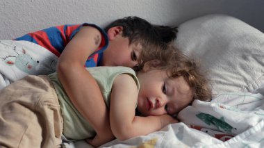 Büyük kardeş küçük kıvırcık saçlı çocuğu yatakta kucaklıyor, uyku sırasında huzur ve koruma anı ya da dinlenme zamanı desenli battaniyenin üzerinde