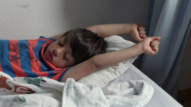 Yatakta uzandıktan sonra kollarını açarak uzanan genç çocuk, uykulu bir ifadeyle ve yumuşak yatak odası ışığında kıvırcık saçlarıyla yavaşça uyanıyor.