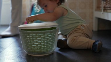 Kıvırcık saçlı meraklı çocuk, mutfak zemininde salata fırıldağıyla oynuyor, erken çocukluk gelişimi sırasında oturmuş ve araştırma yapıyor.