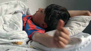 Çocuk yatakta uzanıyor, başı yastığın üzerinde dinleniyor, evde yumuşak bir battaniyenin altında uyukluyor, doğal ışıkla birlikte.
