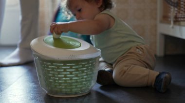 Kıvırcık saçlı meraklı çocuk, mutfak zemininde salata fırıldağıyla oynuyor, erken çocukluk gelişimi sırasında oturmuş ve araştırma yapıyor.