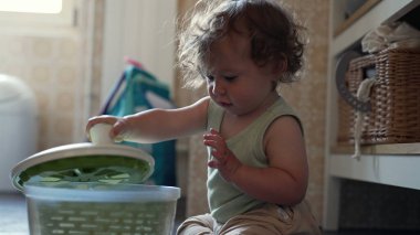 Kıvırcık saçlı meraklı çocuk, mutfak zemininde salata fırıldağıyla oynuyor, erken çocukluk gelişimi sırasında oturmuş ve araştırma yapıyor.