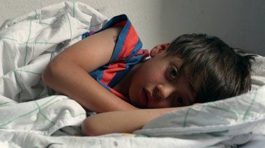 Çocuk yatakta uzanıyor, başı yastığın üzerinde dinleniyor, evde yumuşak bir battaniyenin altında uyukluyor, doğal ışıkla birlikte.