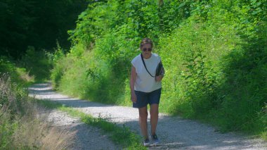 Kıdemli kadın çakıllı orman yolunda yokuş yukarı yürüyor, uzun otlar ve bereketli yaz yeşiliyle çevrili.