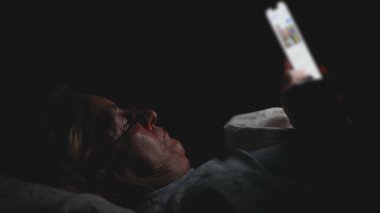 Yaşlı kadın gecenin geç saatlerinde yatakta akıllı telefona bakıyor, karanlık odada içerikte kayarken ekran ışığıyla aydınlatılıyor.
