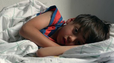 Çocuk yatakta uzanıyor, başı yastığın üzerinde dinleniyor, evde yumuşak bir battaniyenin altında uyukluyor, doğal ışıkla birlikte.