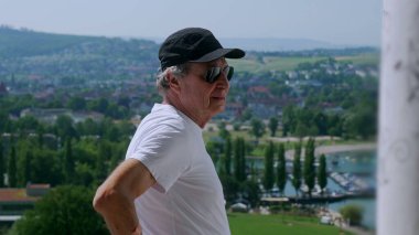 Yaşlı adam tek koluyla dikiliyor. Arka planda yeşil tepeler ve tekneler olan manzaralı bir kasabaya bakıyor.