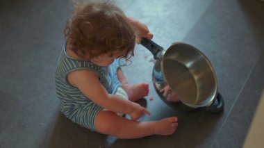 Mutfak zemininde oturmuş yemek pişirme ekipmanlarıyla yaratıcı bir oyun oynarken elinde metal tutacağı tutan bir bebek resmi.