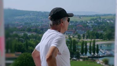 Yaşlı adam tek koluyla dikiliyor. Arka planda yeşil tepeler ve tekneler olan manzaralı bir kasabaya bakıyor.