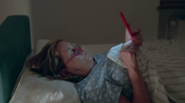 Yaşlı bir kadın, iki eliyle akıllı telefon kullanarak yatakta uzanıyor. Usulca aydınlatılmış odada uyumadan önce ekranla kesişiyor ve etkileşime geçiyor.