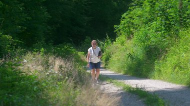 Kıdemli kadın çakıllı orman yolunda yokuş yukarı yürüyor, uzun otlar ve bereketli yaz yeşiliyle çevrili.