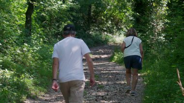 Yaşlı çift birlikte orman yolunda yokuş yukarı yürüyor, ikisi de yeşil ağaçlar ve benekli ışıklarla çevriliyken günlük yaz kıyafetleri giyiyorlar.
