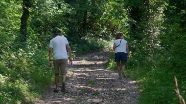 Yaşlı çift birlikte orman yolunda yokuş yukarı yürüyor, ikisi de yeşil ağaçlar ve benekli ışıklarla çevriliyken günlük yaz kıyafetleri giyiyorlar.