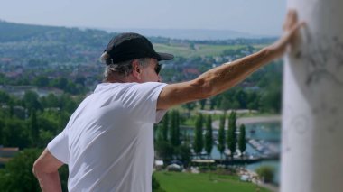 Yaşlı adam tek koluyla dikiliyor. Arka planda yeşil tepeler ve tekneler olan manzaralı bir kasabaya bakıyor.