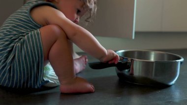 Evde yemek takımlarını keşfederken çıplak ayakla mutfak zemininde otla oynayan meraklı çocuk.