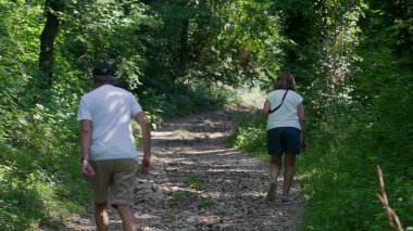 Yaşlı çift birlikte orman yolunda yokuş yukarı yürüyor, ikisi de yeşil ağaçlar ve benekli ışıklarla çevriliyken günlük yaz kıyafetleri giyiyorlar.