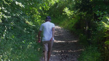 Yaşlı adam orman yolunda tek başına yürüyor, şort ve beyaz gömlekle yokuş yukarı yürüyor, yoğun yeşillik ve sessiz ormanlık atmosferle çevrili.