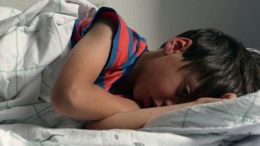 Çocuk yatakta uzanıyor, başı yastığın üzerinde dinleniyor, evde yumuşak bir battaniyenin altında uyukluyor, doğal ışıkla birlikte.