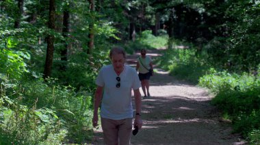 Yaşlı adam gölgeli orman yolunda kadının önünde yürüyor, her iki son sınıf öğrencisi de yemyeşil ormanlarda huzurlu doğa yürüyüşü sırasında yavaşça yokuş yukarı tırmanıyor.