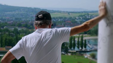 Yaşlı adam tek koluyla dikiliyor. Arka planda yeşil tepeler ve tekneler olan manzaralı bir kasabaya bakıyor.