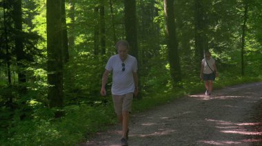 Kıdemli çift orman gölgesinde birlikte yürüyüş yapıyor, erkek önde, kadın arkada, huzurlu ormanlık alanda sessiz bir arkadaşlık.