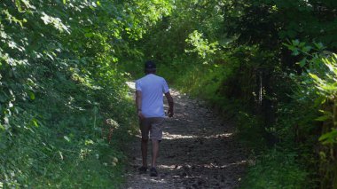 Yaşlı adam orman yolunda tek başına yürüyor, şort ve beyaz gömlekle yokuş yukarı yürüyor, yoğun yeşillik ve sessiz ormanlık atmosferle çevrili.