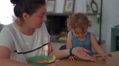 Küçük çocuk çatalla makarna yerken anne izliyor, her ikisi de günlük ev ortamında, parlak doğal ışıkla yemek masasında oturuyor.