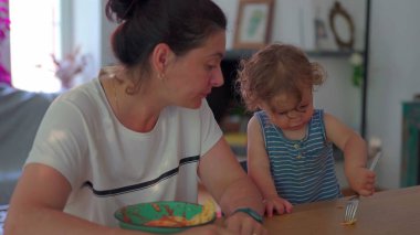 Küçük çocuk çatalla makarna yerken anne izliyor, her ikisi de günlük ev ortamında, parlak doğal ışıkla yemek masasında oturuyor.