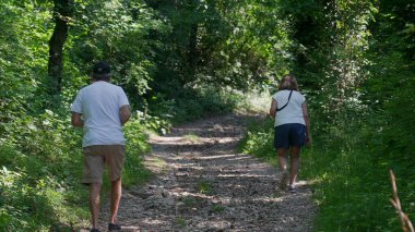 Yaşlı çift birlikte orman yolunda yokuş yukarı yürüyor, ikisi de yeşil ağaçlar ve benekli ışıklarla çevriliyken günlük yaz kıyafetleri giyiyorlar.