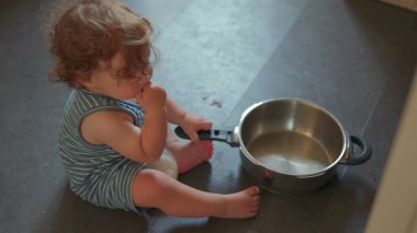 Mutfak zemininde oturmuş yemek pişirme ekipmanlarıyla yaratıcı bir oyun oynarken elinde metal tutacağı tutan bir bebek resmi.