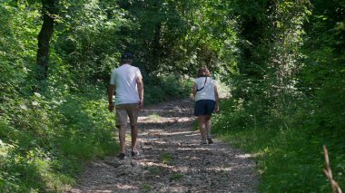 Yaşlı çift birlikte orman yolunda yokuş yukarı yürüyor, ikisi de yeşil ağaçlar ve benekli ışıklarla çevriliyken günlük yaz kıyafetleri giyiyorlar.