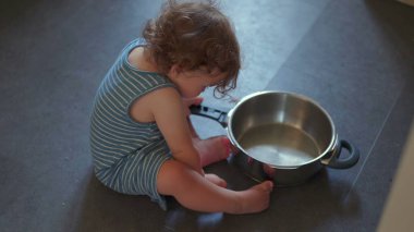 Mutfak zemininde oturmuş yemek pişirme ekipmanlarıyla yaratıcı bir oyun oynarken elinde metal tutacağı tutan bir bebek resmi.