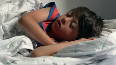 Çocuk yatakta uzanıyor, başı yastığın üzerinde dinleniyor, evde yumuşak bir battaniyenin altında uyukluyor, doğal ışıkla birlikte.