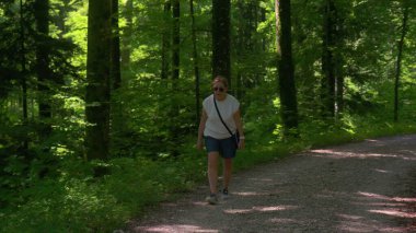 Kıdemli çift orman gölgesinde birlikte yürüyüş yapıyor, erkek önde, kadın arkada, huzurlu ormanlık alanda sessiz bir arkadaşlık.