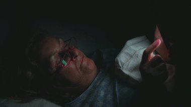 Son sınıf öğrencisi kadın karanlık odada akıllı telefon kullanarak yatakta uzanıyor, gece içeriğinde süzülürken ekranda yansıyan parıltı yüzlere ve gözlüklere yansıyor.