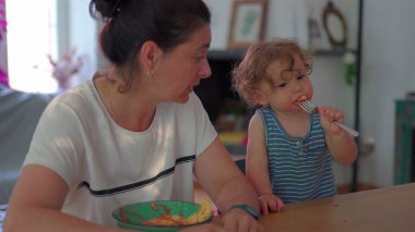Küçük çocuk çatalla makarna yerken anne izliyor, her ikisi de günlük ev ortamında, parlak doğal ışıkla yemek masasında oturuyor.