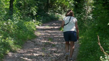 Yaşlı kadın birlikte orman yolunda yokuş yukarı yürüyor, ikisi de yeşil ağaçlar ve benekli ışıklarla çevriliyken günlük yaz kıyafetleri giyiyor.