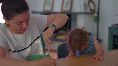 Küçük çocuk çatalla makarna yerken anne izliyor, her ikisi de günlük ev ortamında, parlak doğal ışıkla yemek masasında oturuyor.