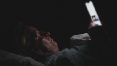 Yaşlı kadın gecenin geç saatlerinde yatakta akıllı telefona bakıyor, karanlık odada içerikte kayarken ekran ışığıyla aydınlatılıyor.