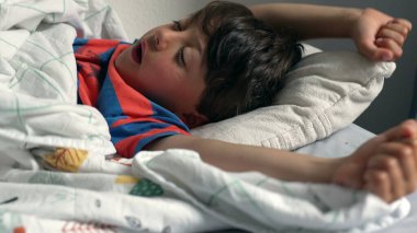 Çocuk yatakta uzanıyor, başı yastığın üzerinde dinleniyor, evde yumuşak bir battaniyenin altında uyukluyor, doğal ışıkla birlikte.