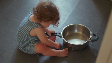 Mutfak zemininde oturmuş yemek pişirme ekipmanlarıyla yaratıcı bir oyun oynarken elinde metal tutacağı tutan bir bebek resmi.